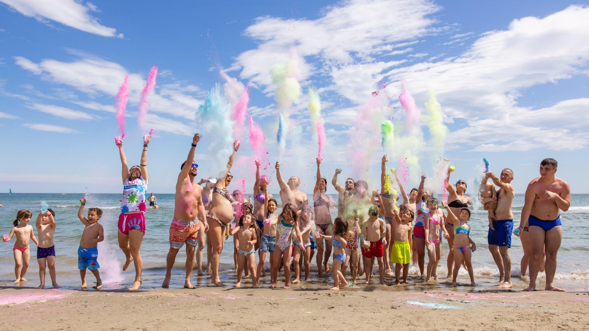 Persone lanciano polveri colorate sulla spiaggia in una giornata soleggiata.