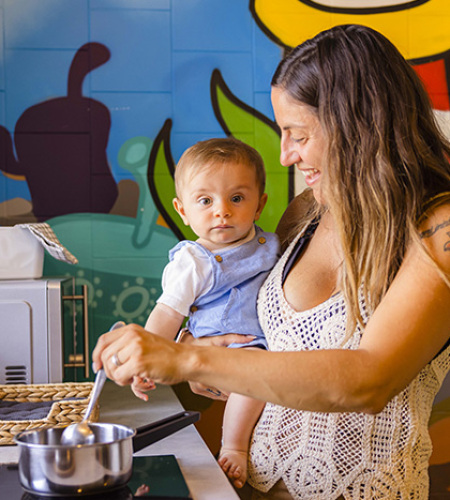 Famiglia in cucina, madre prepara, bambino sorride, altra donna dà da mangiare al piccolo.