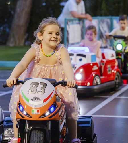 Smiling children on toy motorcycles in a sunny amusement park.