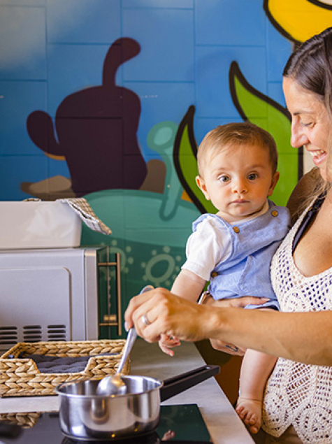Famiglia in cucina, madre prepara, bambino sorride, altra donna dà da mangiare al piccolo.