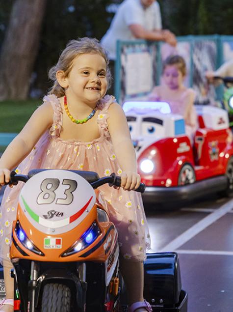 Smiling children on toy motorcycles in a sunny amusement park.