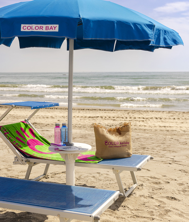 Ragazzo sistema ombrellone su spiaggia deserta, indossando occhiali da sole e maglietta blu.