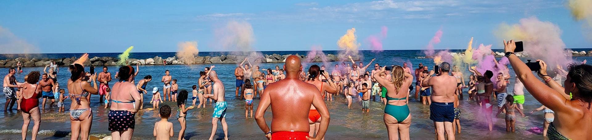 Persone in spiaggia lanciano polveri colorate durante una festa estiva.